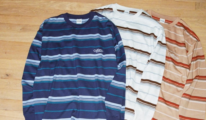 Three California General Store long-sleeve multi-border t-shirts in blue, white, and tan colorways laid flat on a wood floor.