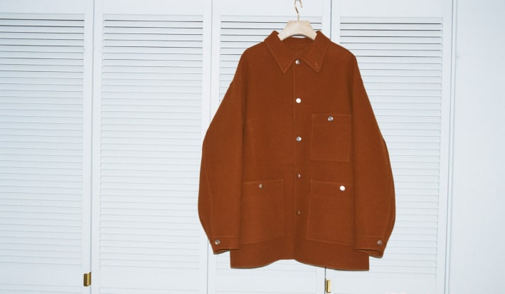 A rust-colored wool chore jacket with silver buttons hangs from a wooden hanger against a white louvered door.